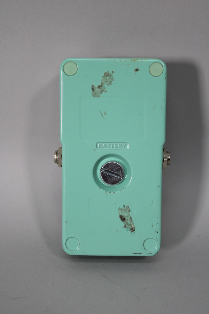 The pedal's bottom is teal with four light green rubber feet, a circular battery compartment, and visible grime and wear.