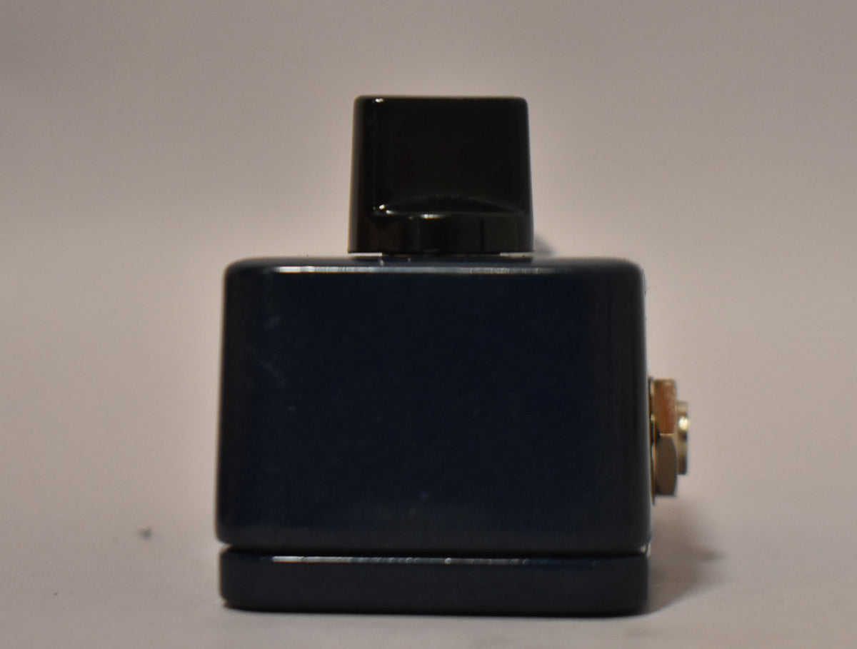 The dark blue pedal is viewed from the side, showing a black footswitch on top and a gold jack on the right.