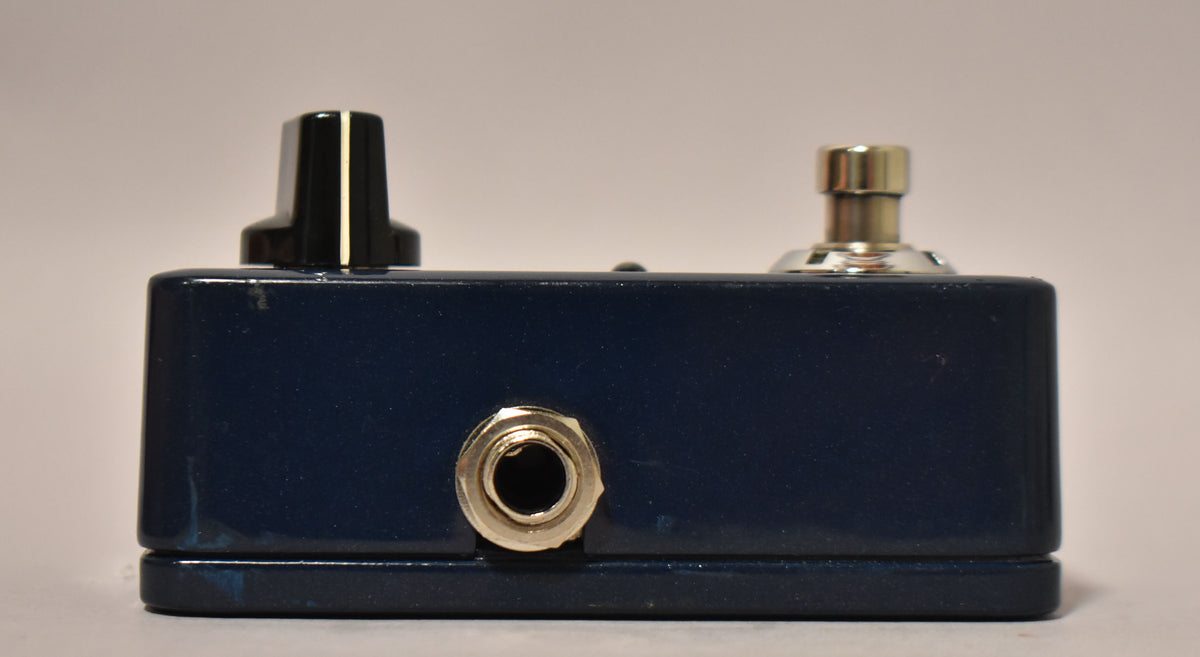 Side view of a dark blue metallic pedal with a silver input jack, a black knob, and a silver footswitch.
