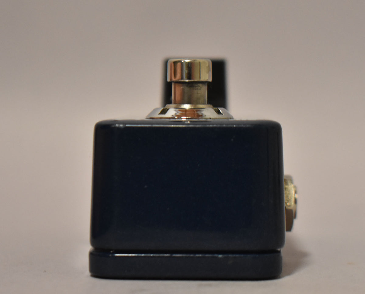 A dark blue guitar pedal with a silver footswitch and a gold jack on the side, against a light gray background.