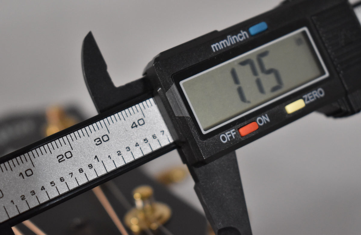 A digital caliper measures the guitar's nut width at 1.75 inches, with gold tuning pegs visible in the background.