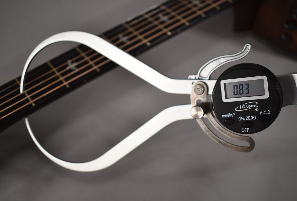 A digital caliper measures the guitar's fretboard, displaying "0.83" on its screen, with the neck angled from left to right.