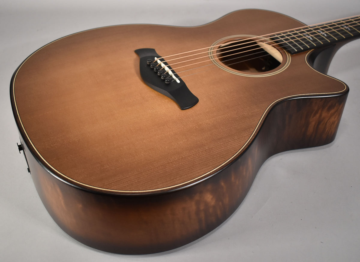The acoustic guitar's body, with a matte honey burst top and dark figured sides, is angled from the lower bout.
