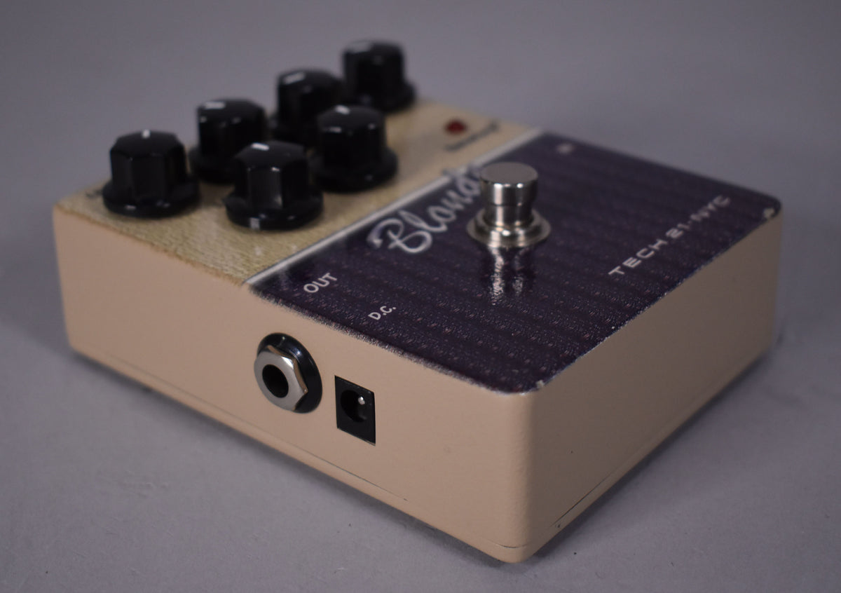 The pedal is angled from the left, showing the beige side with an output jack and power input, and the dark purple top with six black knobs and a silver footswitch.