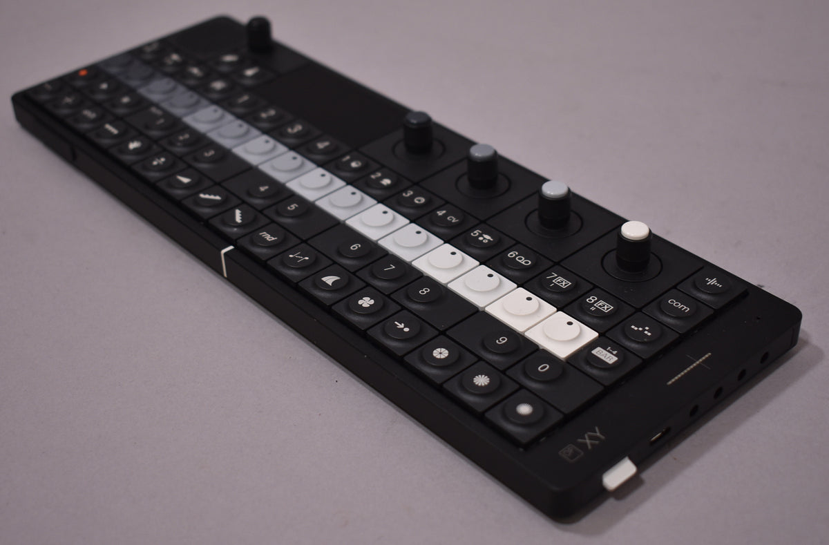 The black synthesizer is angled from above, showing its full length, with a row of white and gray buttons and four knobs.