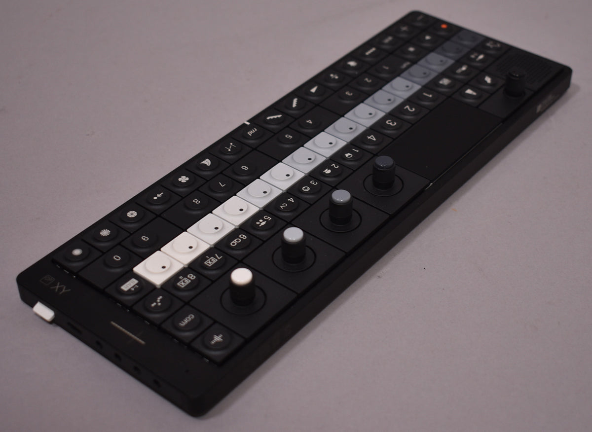 Black synthesizer, angled from the left, showing a grid of black, white, and gray buttons, plus four joysticks.