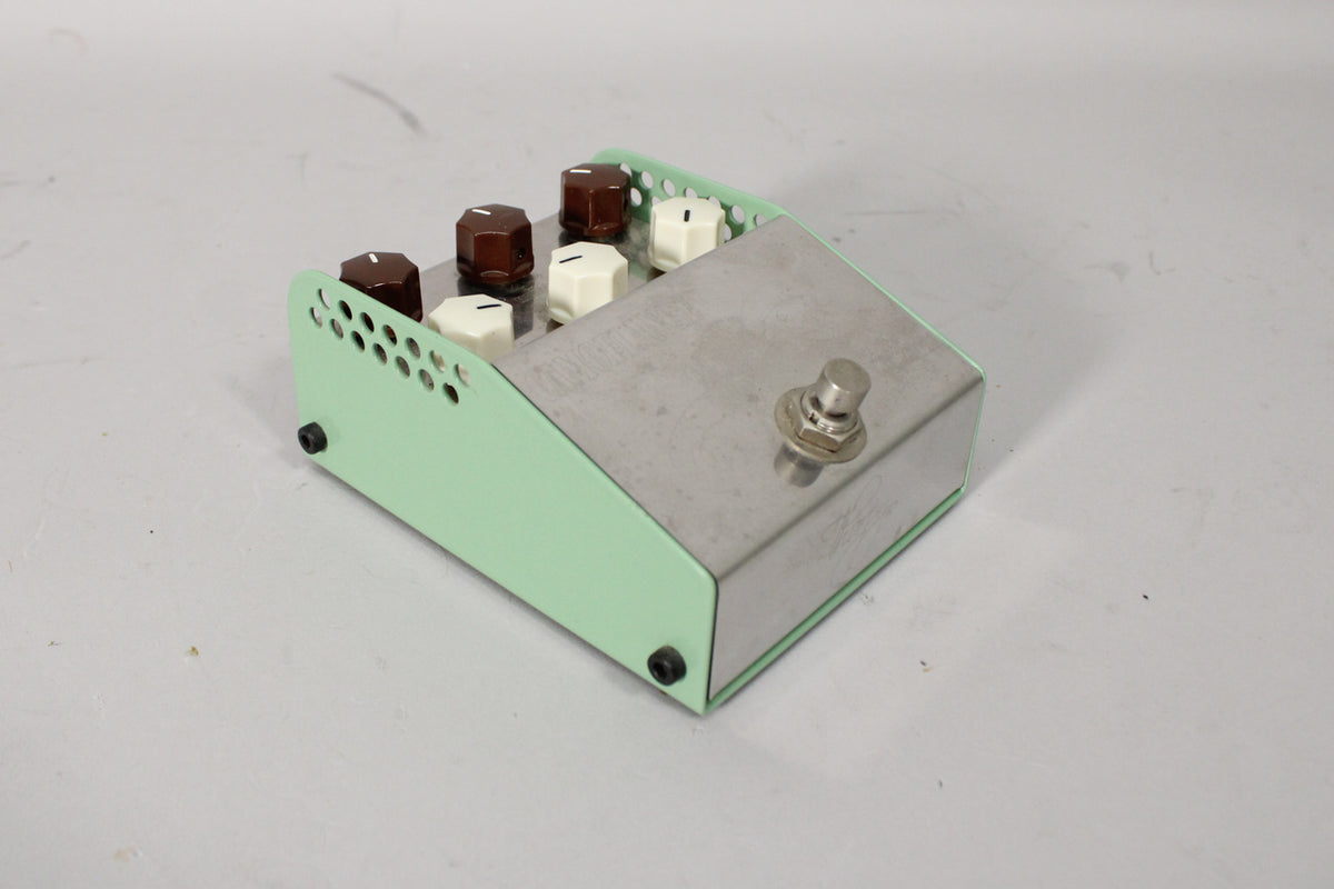 Angled view of the mint green and brushed metal pedal, showing six control knobs, three brown and three cream, and a footswitch.