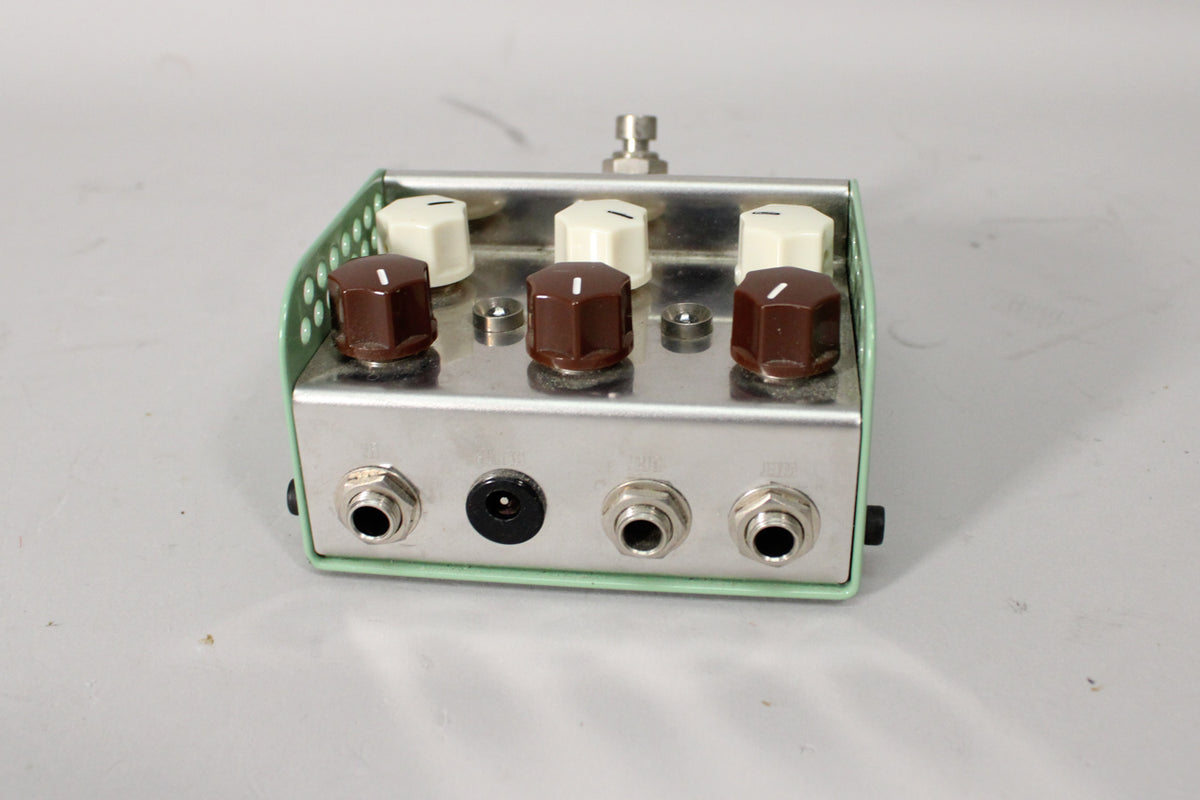 A light green and silver guitar pedal with six knobs, three brown and three cream, and four input jacks.