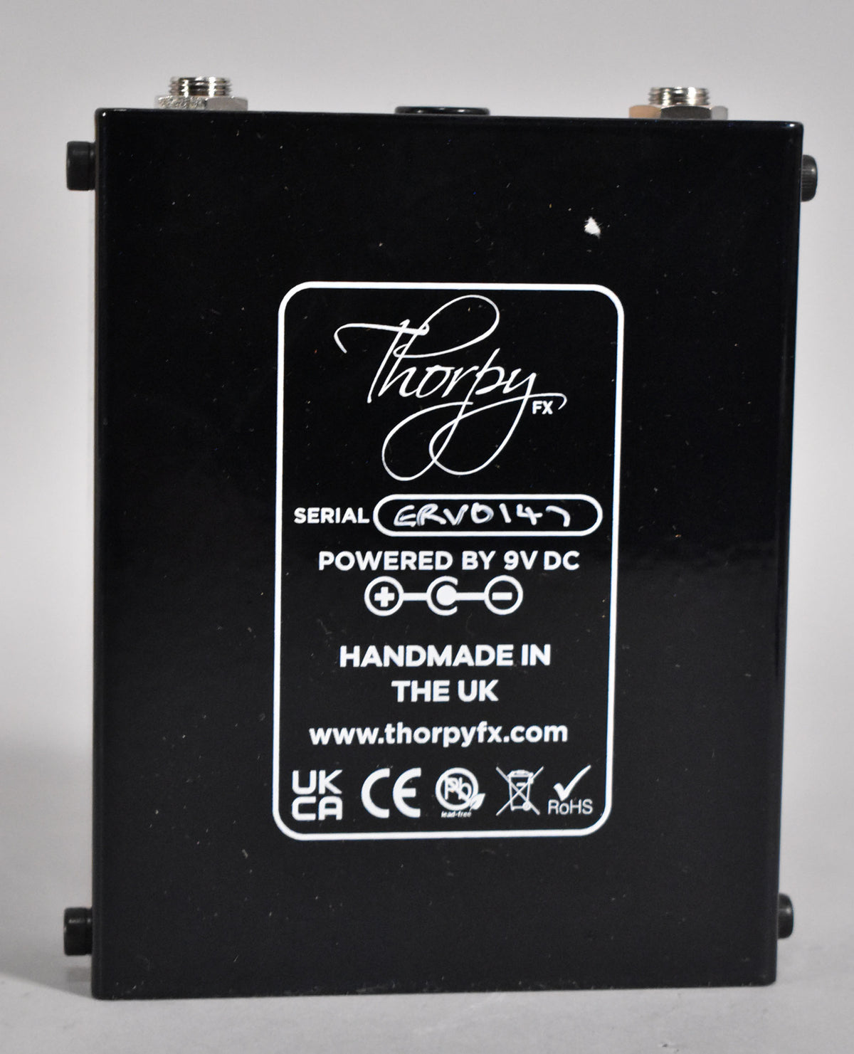 The black pedal's back panel is visible, with a white label detailing serial number, 9V DC power, and UK manufacturing.