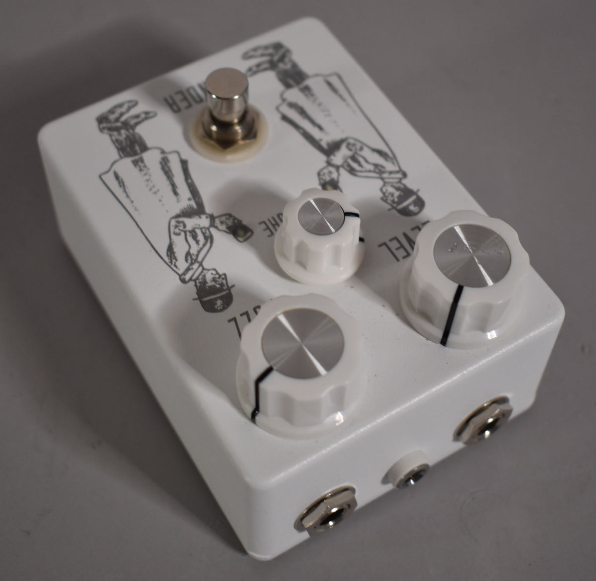 White pedal with black line art of two figures, a silver footswitch, three white knobs with silver caps, and two input jacks.