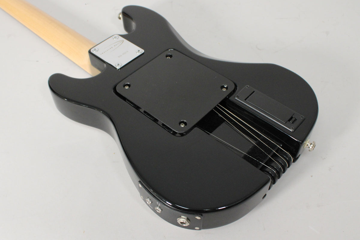 The guitar's glossy black body is seen from the back, showing a light wood neck, black control plates, and a battery compartment.
