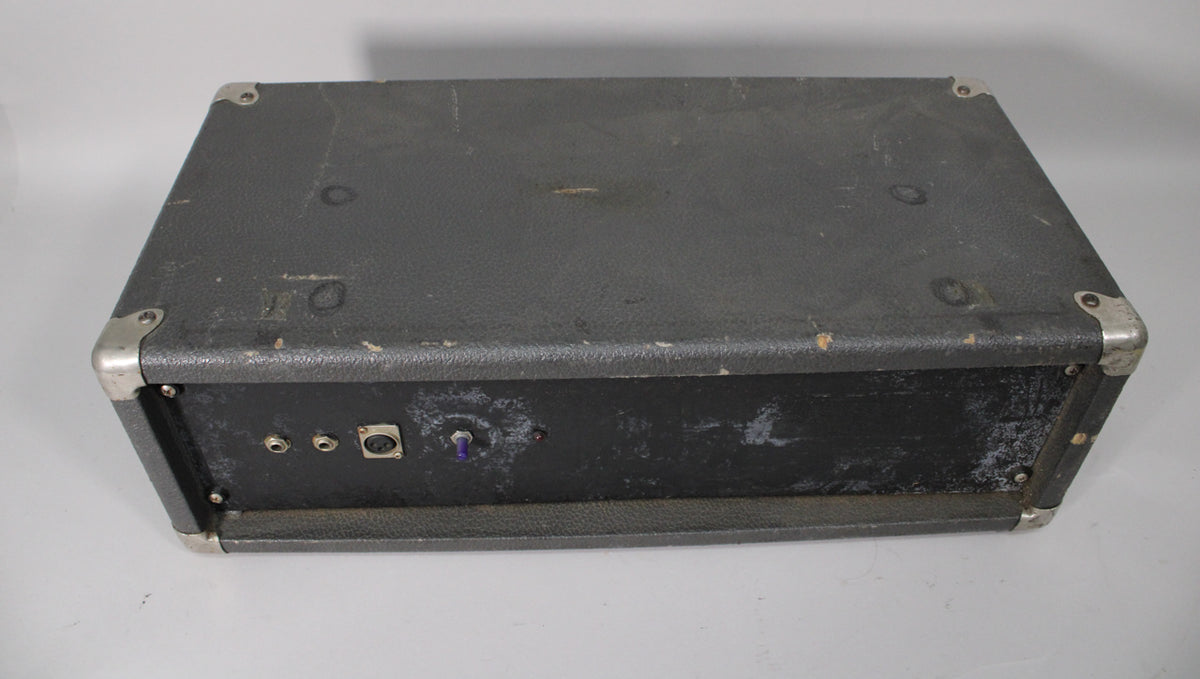 Top-down view of a dark gray amplifier with a textured finish, showing the back panel with three jacks and a purple toggle switch.