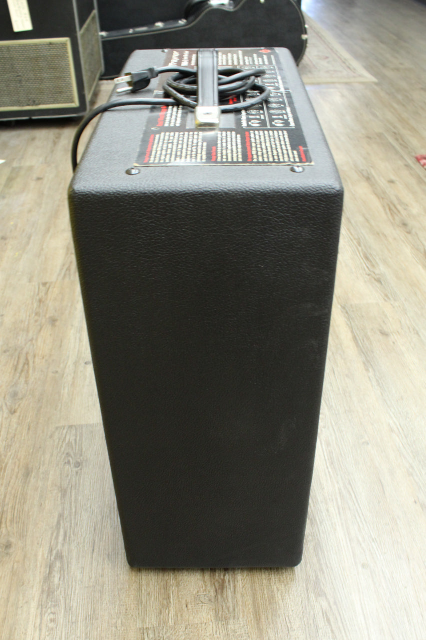 The black textured amp is seen from above, showing its top panel with a handle, coiled power cord, and information labels.