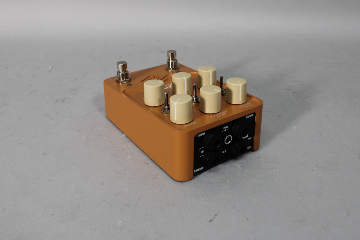 An orange guitar pedal with six cream-colored knobs, three toggle switches, and various input/output jacks on a gray surface.