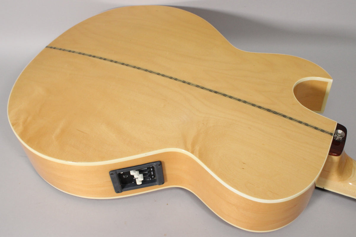 The natural finish acoustic bass's back and side are visible, showing a dark center strip and black preamp controls on the lower bout.