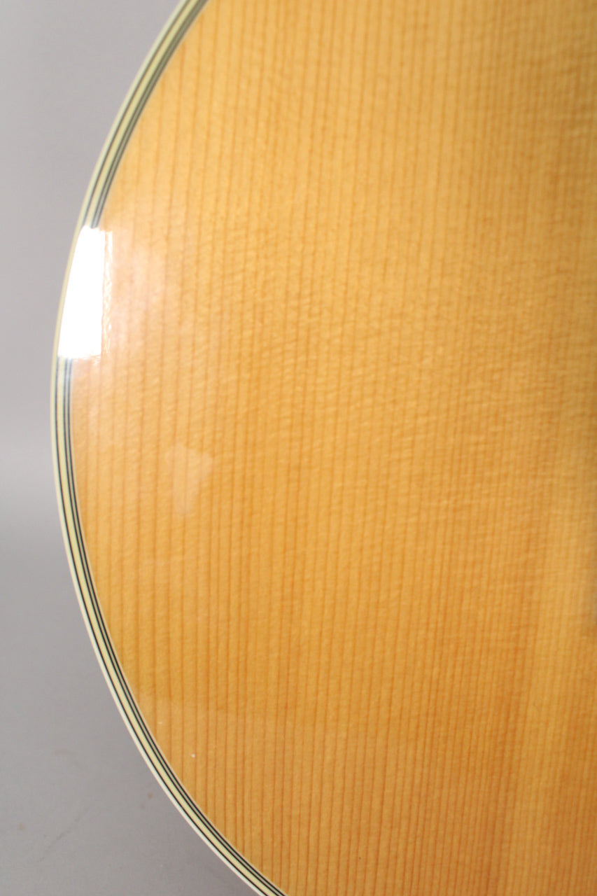 Close-up of the bass's natural wood top, showing fine vertical grain, a glossy finish, and multi-ply binding along the curved edge.