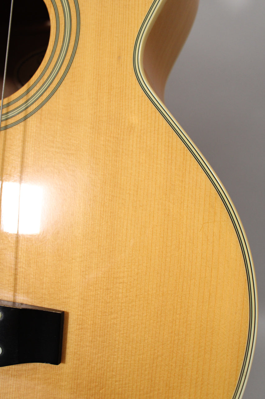 Close-up of the natural finish acoustic bass body, showing the soundhole, black bridge, and multi-line binding.