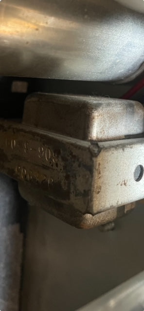 Close-up of a tarnished, rectangular silver-colored metal component with faint numbers stamped on its side, showing wear and rust.