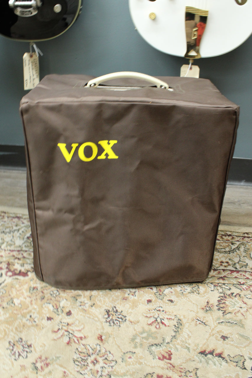 The amp is covered by a brown fabric cover with a yellow logo, showing a cream handle on top.