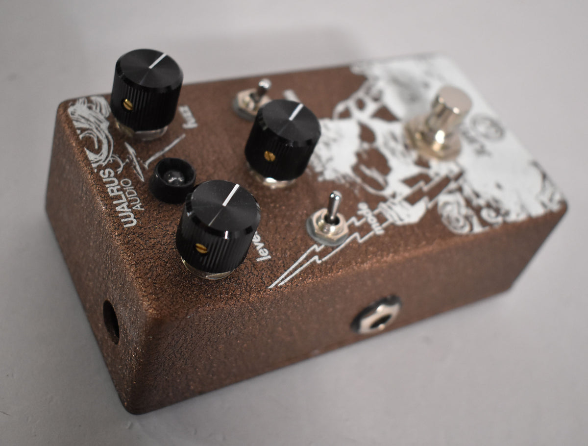 A top-down view of a textured brown pedal with white skull graphics, three black knobs, two toggle switches, and a footswitch.
