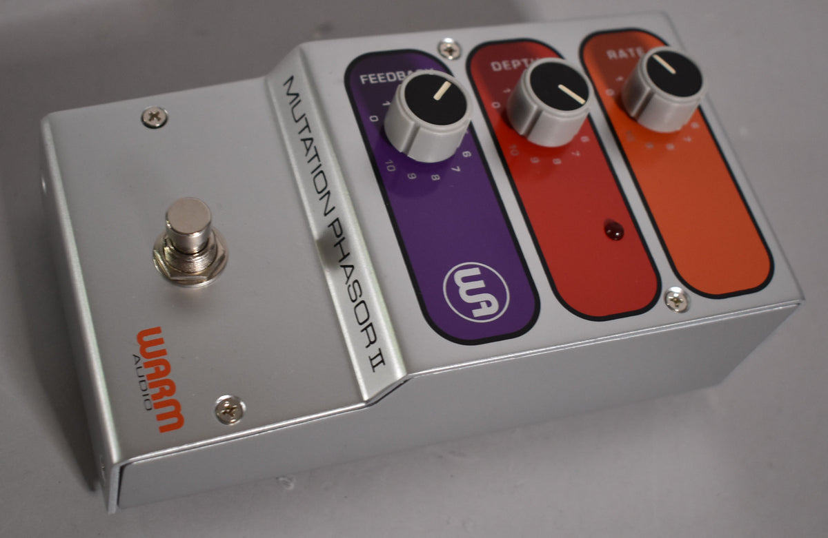A silver guitar pedal, viewed from above and the right, with a footswitch and three black-capped knobs over purple, red, and orange panels.