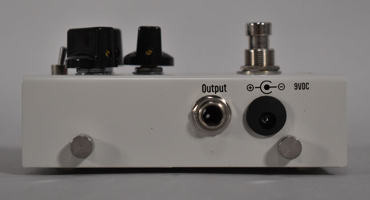 The white pedal is viewed from the rear, showing the output jack, 9VDC power input, and two black control knobs on top.