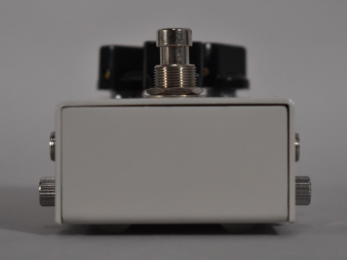 Front view of a white guitar pedal with a silver footswitch and two black knobs visible in the background.