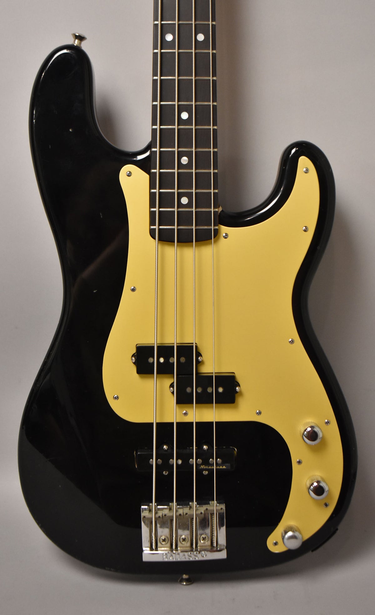 Close-up of a glossy black bass with a cream pickguard, showing the neck, PJ pickups, bridge, and three chrome knobs.