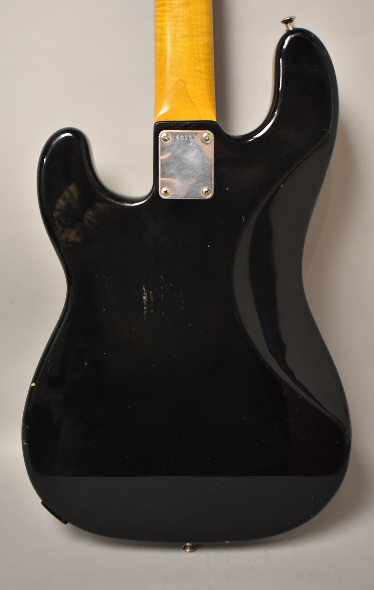 Rear view of a black PJ-style bass body with a maple neck, showing a worn neck plate and many scratches on the glossy finish.