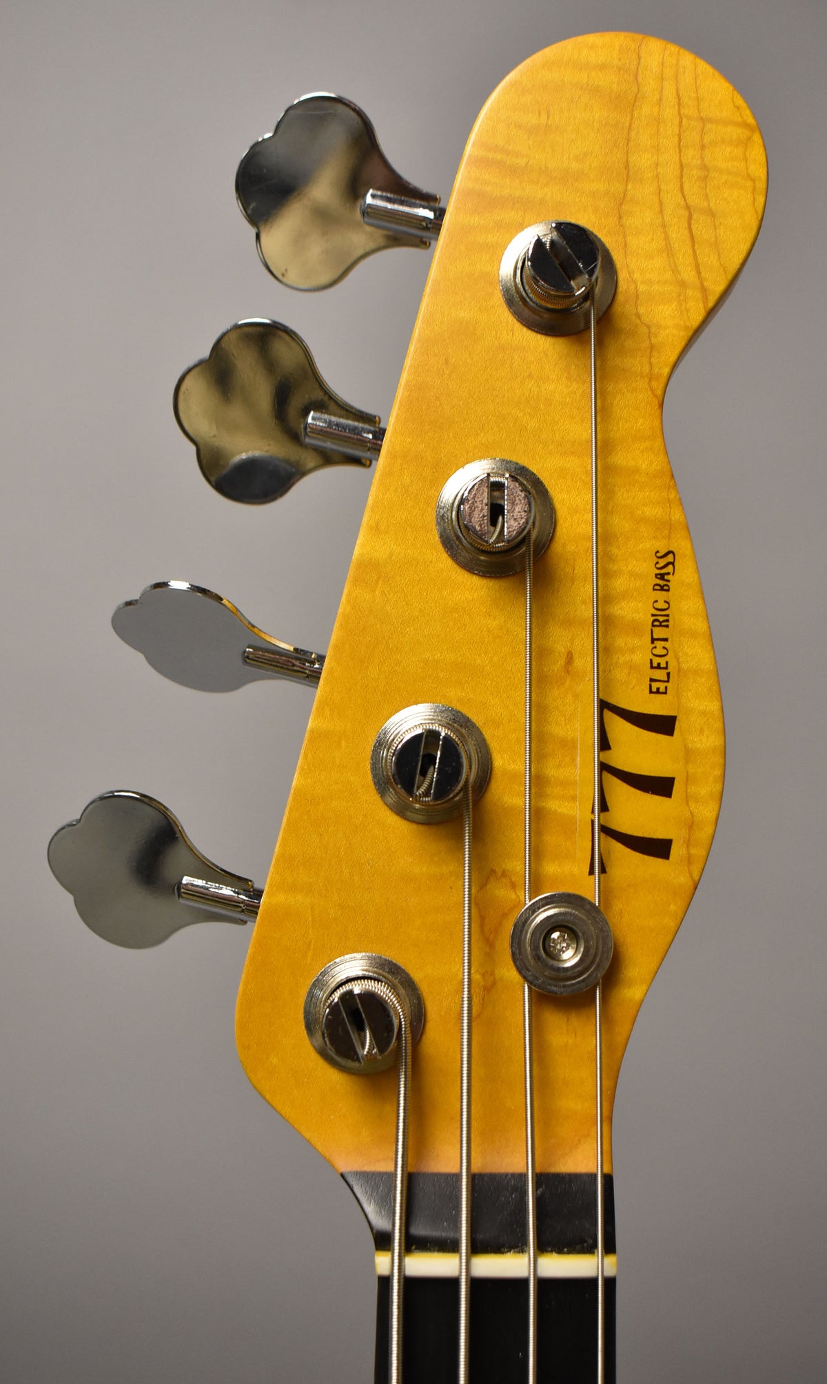 Close-up of a bass headstock, angled right, showing the amber-finished maple with four chrome clover-leaf tuners and string tree.