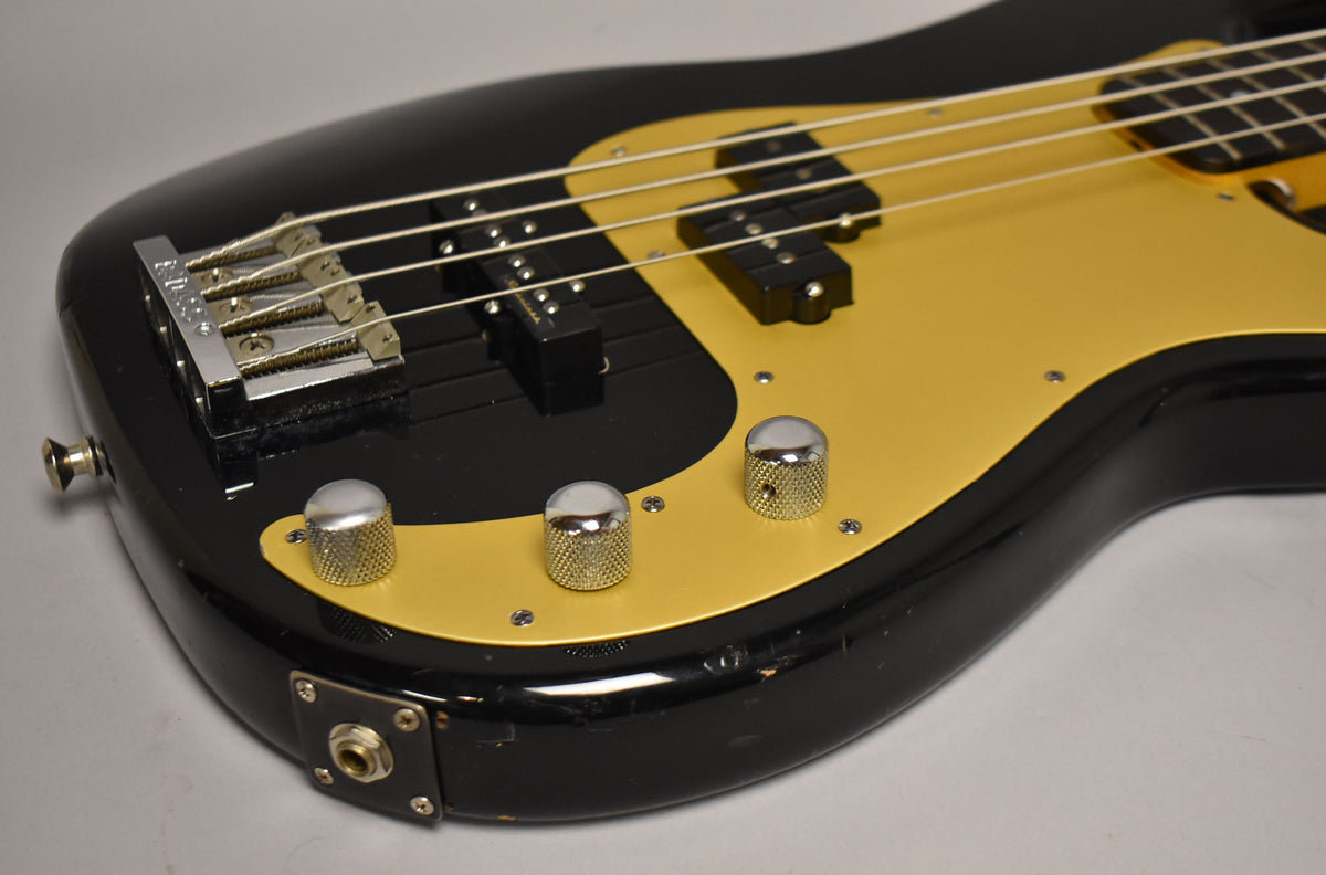 Close-up of a black bass body with a gold pickguard, chrome bridge, two pickups, three knurled chrome knobs, and a worn output jack.