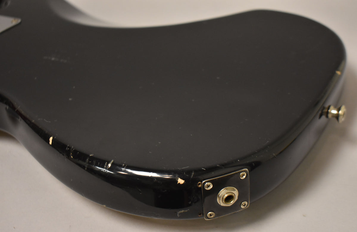 The lower bout of a black bass body, showing the input jack and strap button, with visible chips and scratches.