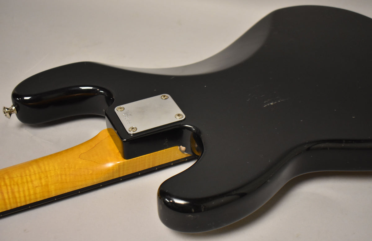 Rear view of a black bass body with a maple neck, showing a chrome neck plate and a chip in the finish near the neck joint.