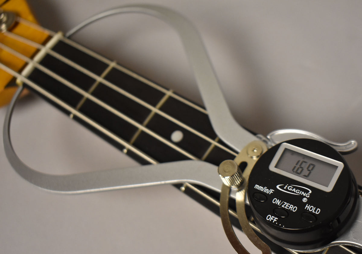 Close-up of the bass neck with a digital caliper measuring the nut width at 1.69 inches, showing the dark fretboard and light frets.