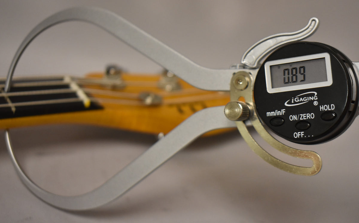 A digital caliper measures the bass neck's thickness at the nut, showing "0.89" on its display.