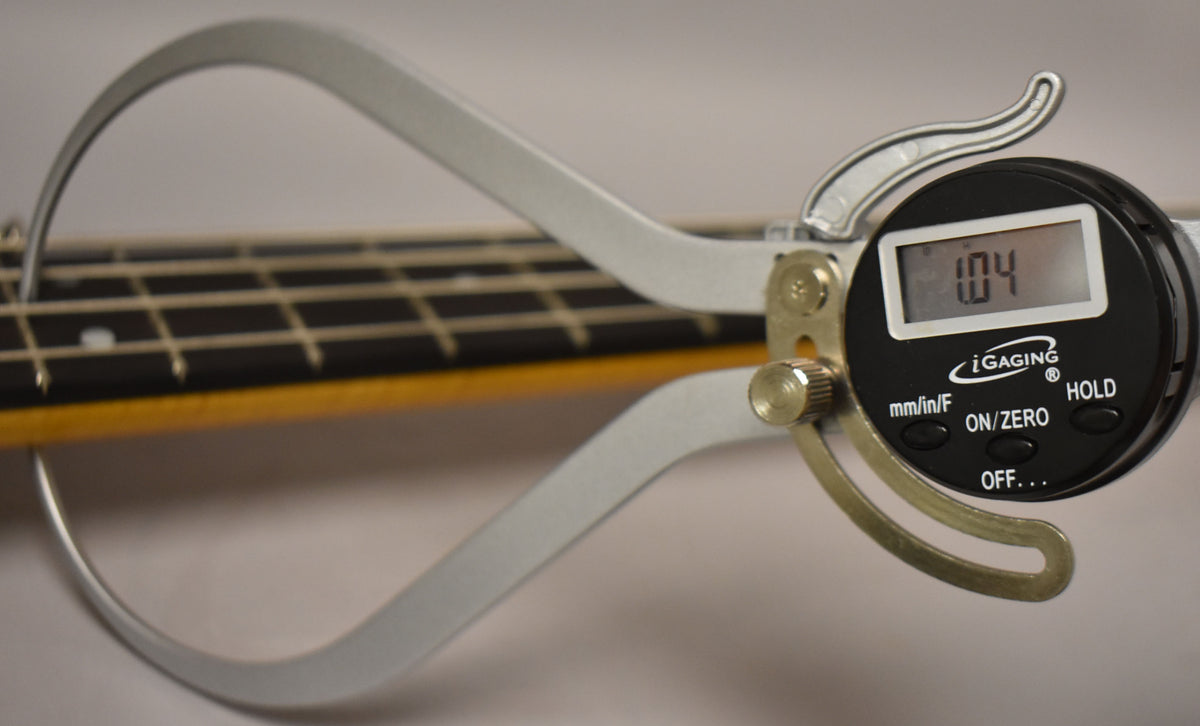 A digital caliper measures the bass's fretboard thickness, displaying "1.04" on its screen, with the neck angled from lower left.