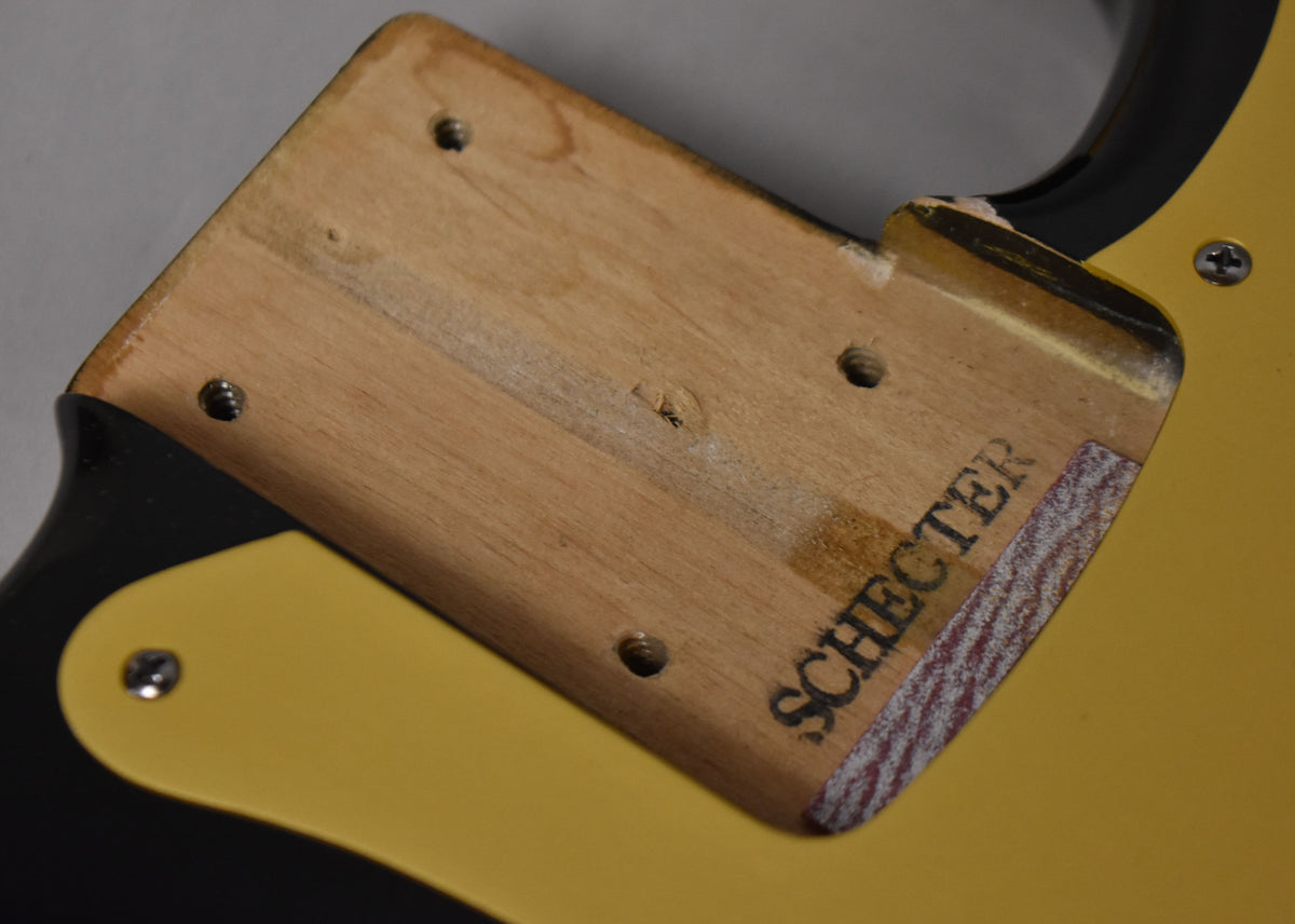 Close-up of the bass's neck pocket, showing exposed wood, four screw holes, and a "Schecter" stamp on the body.