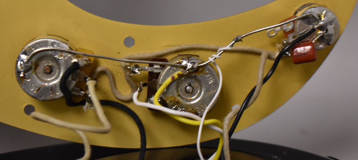 Close-up of the bass's control cavity, showing three tarnished pots, a brown capacitor, and vintage-style cloth wiring on a yellow pickguard.