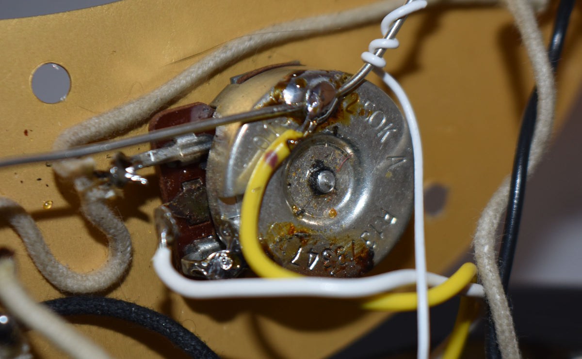 Close-up of the bass's internal wiring, showing a tarnished silver potentiometer with yellow and white wires soldered to it.