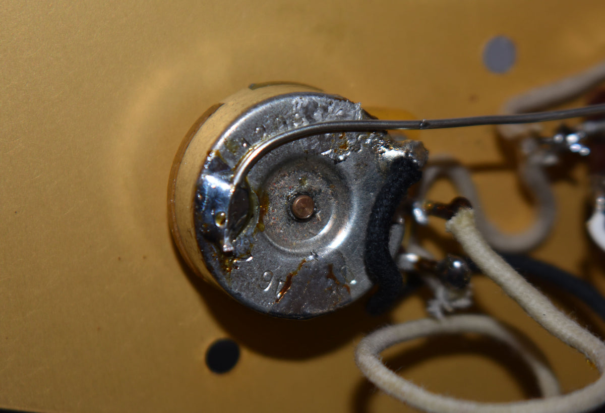 Close-up of a tarnished chrome potentiometer with a copper shaft, showing solder joints and cloth-wrapped wires against a gold surface.