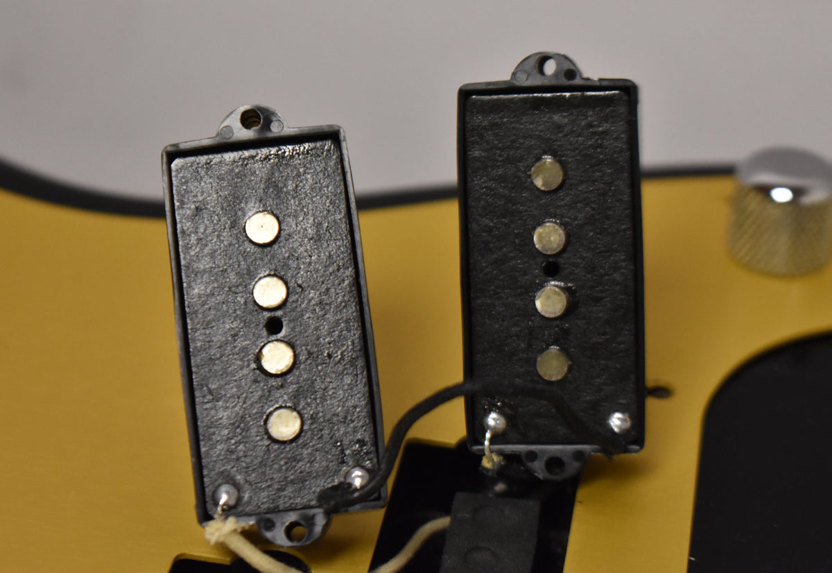 Close-up of two black PJ-style bass pickups with gold pole pieces, showing wiring and a chrome control knob on a yellow pickguard.