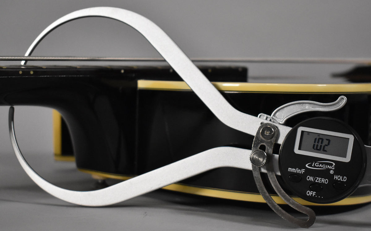A digital caliper measures the string height above the frets on the bass's black neck, showing "1.02" mm.