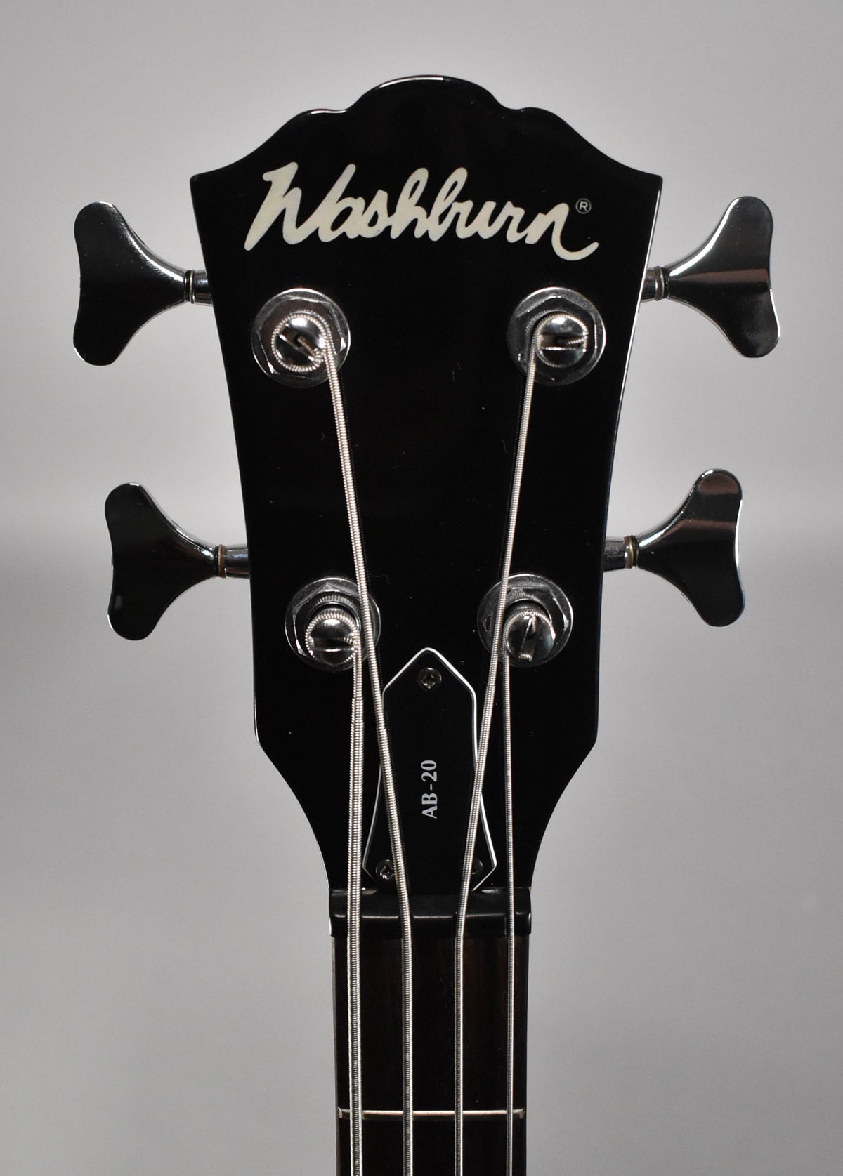 Close-up of the bass's glossy black headstock, showing four chrome tuning pegs, strings, and a black truss rod cover.