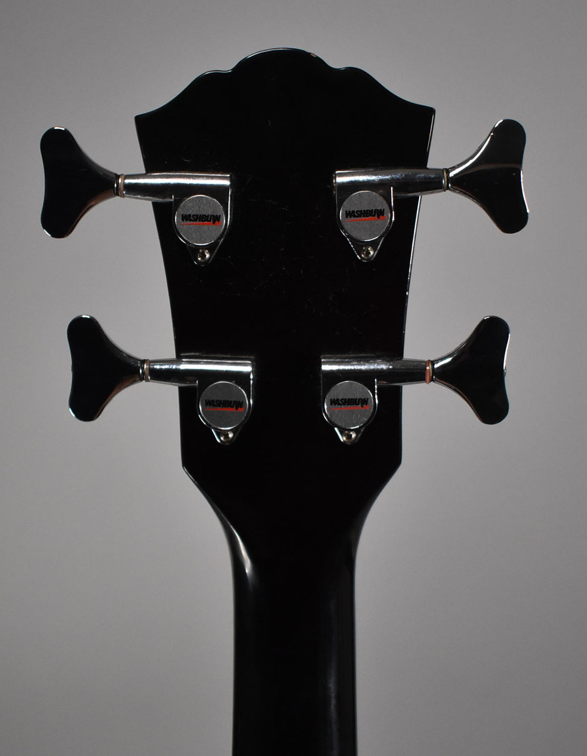 The bass's glossy black headstock and neck, viewed from behind, show four chrome tuners with black paddle keys.