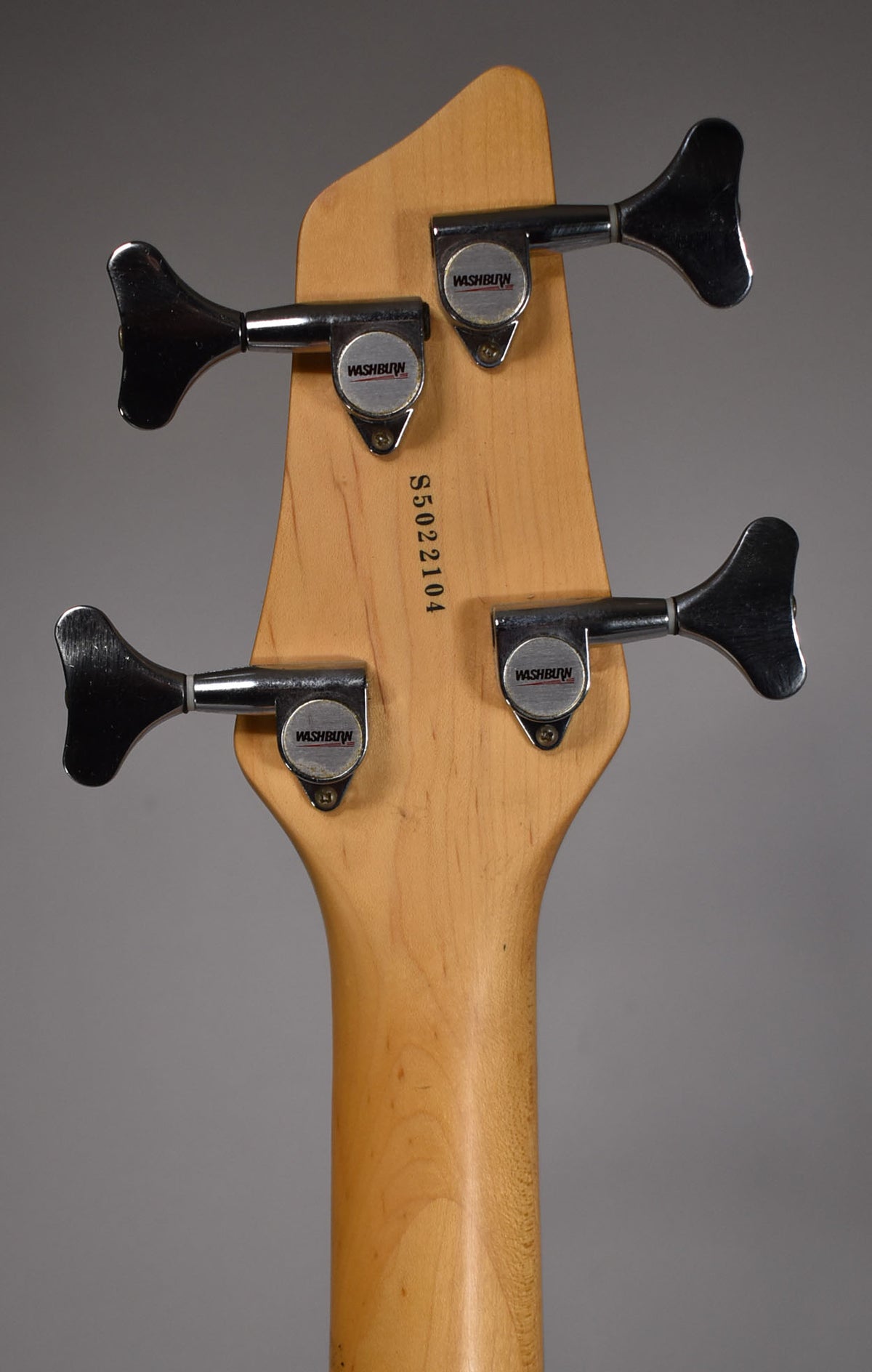 Rear view of the bass headstock and upper neck, showing four chrome tuners on a natural wood finish, with some light wear.