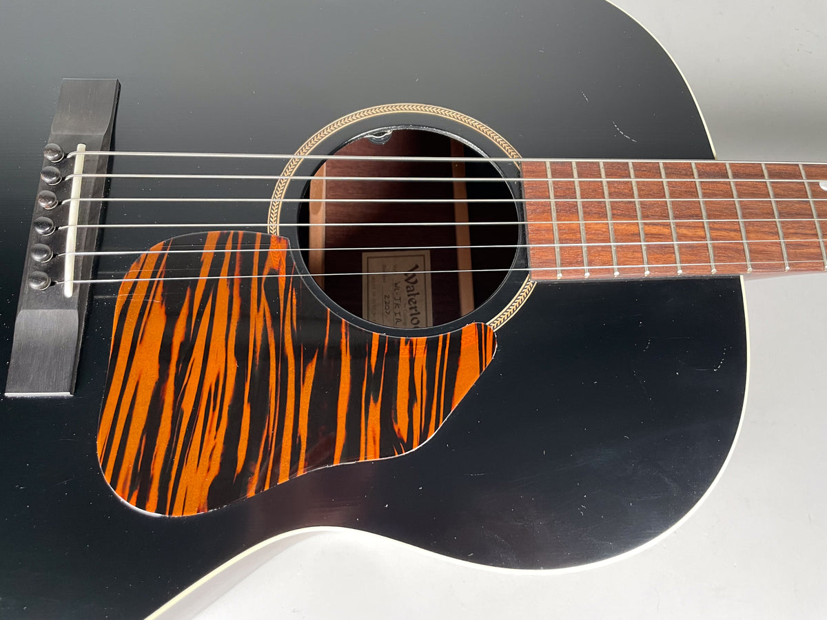 Close-up of the black acoustic guitar's body, showing a tiger-stripe pickguard, soundhole rosette, and light wear on the finish.