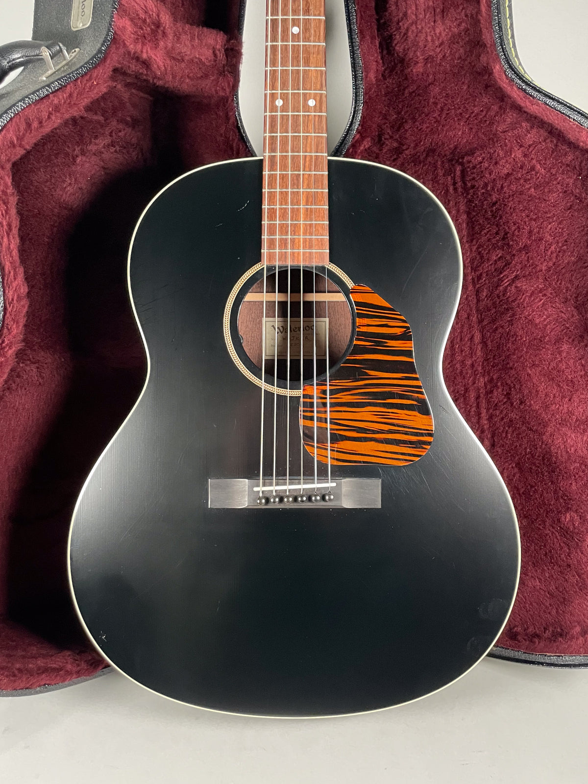 The black acoustic guitar is angled slightly, showing its body, fretboard, and a tiger-stripe pickguard, resting in a red plush case.