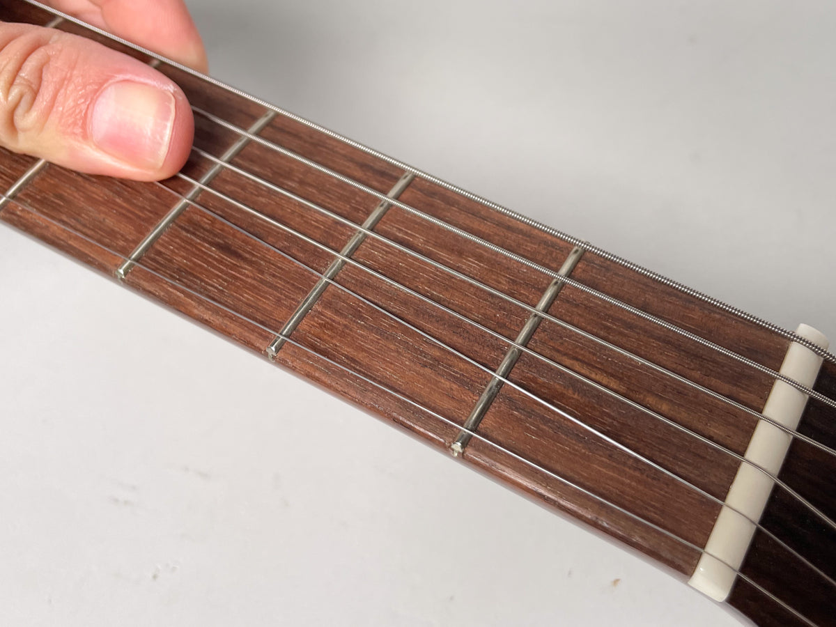 Close-up of the dark brown fretboard and strings, with a thumb pressing down on the second fret.