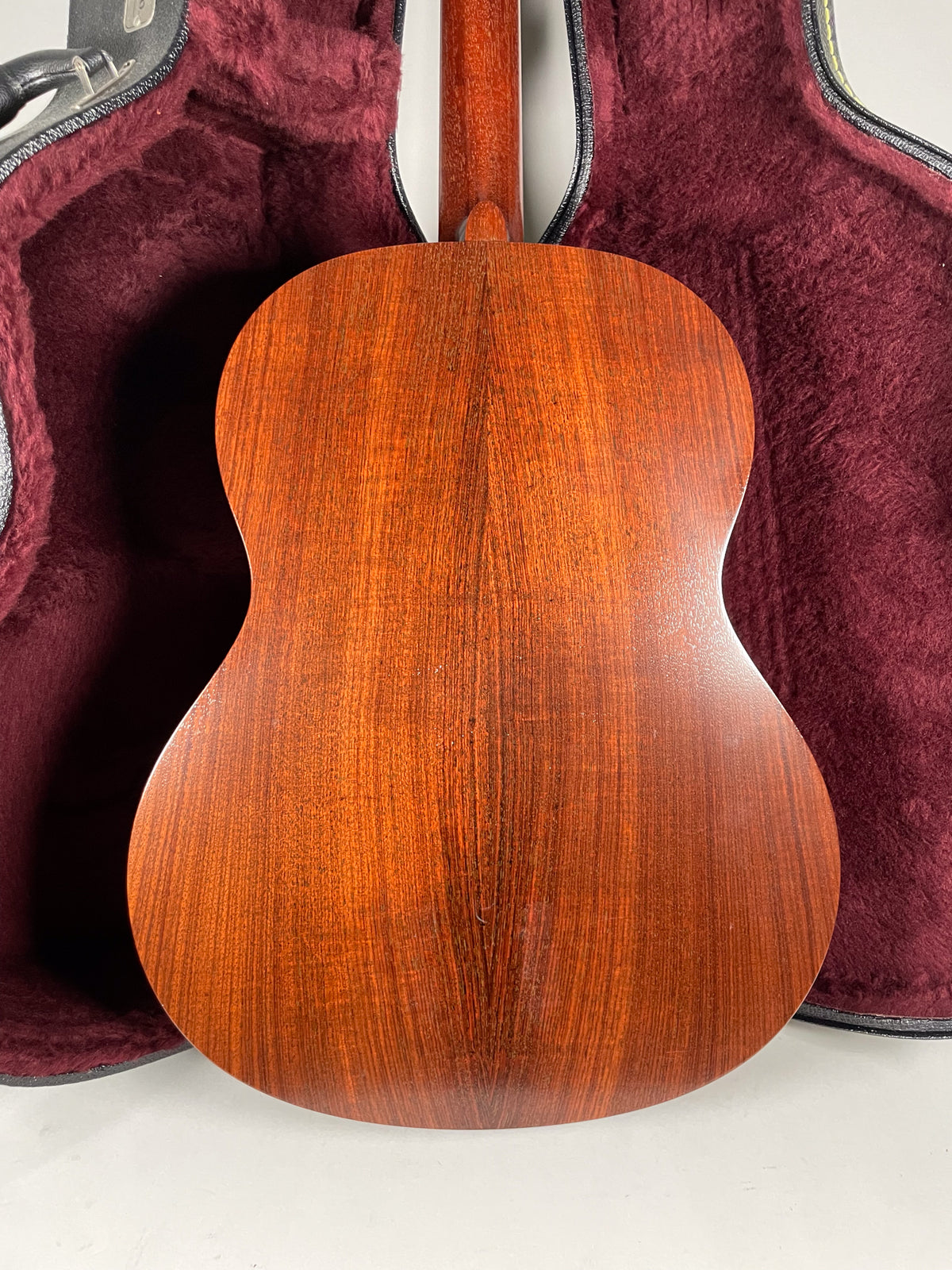 The back of the acoustic guitar, with a rich reddish-brown wood grain and a visible seam down the center, rests in a burgundy case.