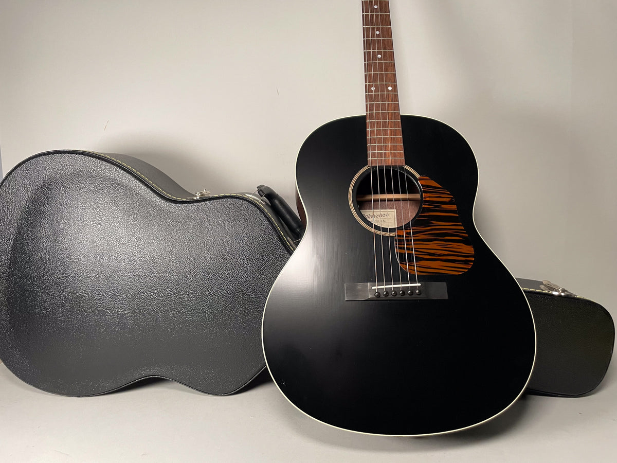 A black acoustic guitar with a matte finish and a tiger-stripe pickguard stands next to its open hard case.
